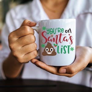May include: White ceramic mug with a green, red and white design that says "You're on Santa's List" with a brown poop emoji with a smiley face.