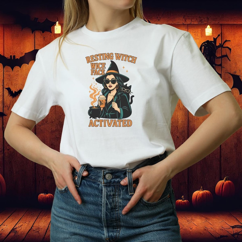May include: White t-shirt with a Halloween-themed graphic. The design features a witch with sunglasses, a black cat, and the text "Resting Witch Face Activated" in orange. The witch is holding a cup of coffee.