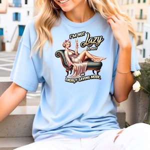 Puede incluir: Camiseta azul claro con una ilustraci&oacute;n de dibujos animados de una mujer tumbada en una chaise longue, sosteniendo una copa de champ&aacute;n. El texto de la camiseta dice "I'M NOT Lazy, I'M JUST ON ENERGY-SAVING MODE."