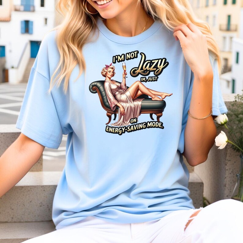 Puede incluir: Camiseta azul claro con una ilustraci&oacute;n de dibujos animados de una mujer tumbada en una chaise longue, sosteniendo una copa de champ&aacute;n. El texto de la camiseta dice "I'M NOT Lazy, I'M JUST ON ENERGY-SAVING MODE."