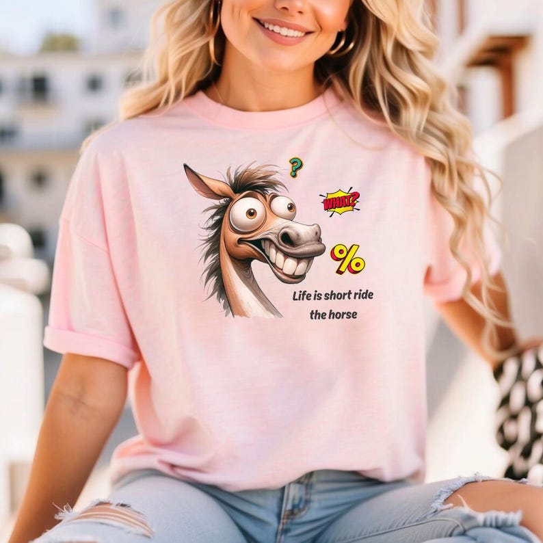 May include: A light pink t-shirt featuring a cartoon horse with a large grin and the text "Life is short ride the horse." The horse graphic includes speech bubbles and a question mark.