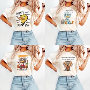 May include: Four cream-colored graphic t-shirts with different cartoon animal designs and text. One shirt features a duck with the text "WHAT? FUCK YOU". Another says "Monday" with a duck wearing glasses. The third says "DINNERT" and the last says "I HAVE NO IDEA WHAT'S GOING ON... BUT I'M HERE FOR IT!"