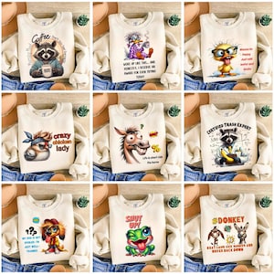 May include: A collection of cream-colored sweatshirts with various cartoon graphics and text. Designs include a raccoon with coffee, a duck in pajamas, a chicken, a horse, a raccoon with a banana, a dog, a frog, and donkeys. The sweatshirts are displayed on a denim background.