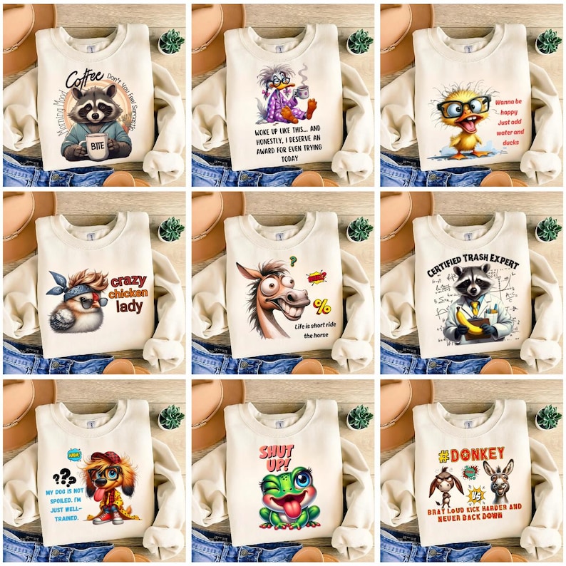 May include: A collection of cream-colored sweatshirts with various cartoon graphics and text. Designs include a raccoon with coffee, a duck in pajamas, a chicken, a horse, a raccoon with a banana, a dog, a frog, and donkeys. The sweatshirts are displayed on a denim background.