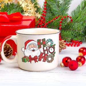 May include: White ceramic mug with a black rim and handle. The mug features a cartoon Santa Claus with a red hat and beard, and the words "Ho Ho" in a colorful, patterned design.