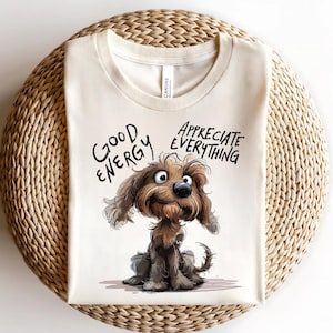 May include: Cream-colored t-shirt featuring a cartoon dog illustration. The dog is brown and has a friendly expression. The shirt has the words "GOOD ENERGY" and "APPRECIATE EVERYTHING" in black script. The shirt is folded on a woven surface.