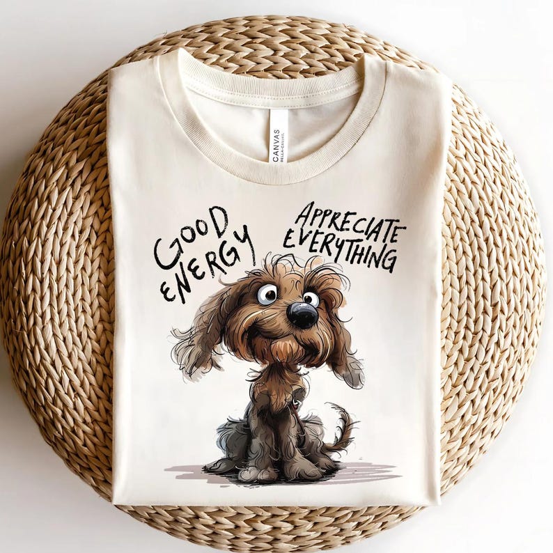 May include: Cream-colored t-shirt featuring a cartoon dog illustration. The dog is brown and has a friendly expression. The shirt has the words "GOOD ENERGY" and "APPRECIATE EVERYTHING" in black script. The shirt is folded on a woven surface.