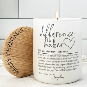 May include: A white candle with the text "difference maker" and a heart symbol, with a definition below. The candle is lit, and the lid, engraved with "MERRY CHRISTMAS", is next to it. A thoughtful gift.