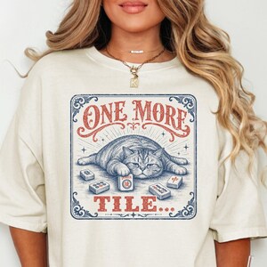 May include: Cream-colored t-shirt featuring a graphic of a cat lying down with the text "ONE MORE TILE...". The cat is surrounded by Mahjong tiles. The shirt is paired with denim shorts.