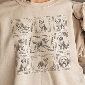 May include: Beige sweatshirt featuring a grid of nine dog illustrations designed to look like postage stamps. The dogs are depicted in various poses, with a vintage aesthetic. The sweatshirt has a crew neck and long sleeves.