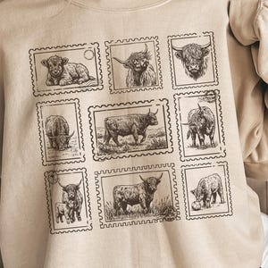 May include: Beige sweatshirt featuring a grid of nine Highland cow illustrations within postage stamp frames. The black and white drawings depict various poses of the cows. The sweatshirt has a crew neck and long sleeves.