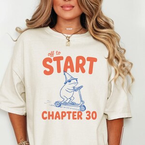 May include: Cream-colored t-shirt with the text "off to START CHAPTER 30" in orange and blue. The graphic features a frog riding a scooter. The shirt is worn with denim shorts.