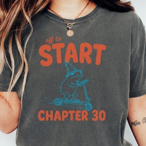 May include: Dark gray t-shirt with the text "off to START CHAPTER 30" in orange and a blue illustration of a frog on a scooter. The shirt is a casual, unisex style.