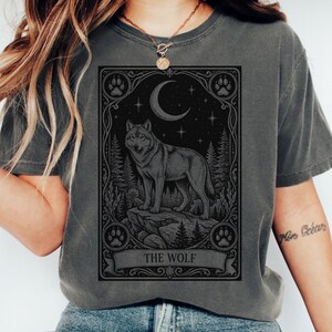 May include: A charcoal gray t-shirt featuring a rectangular graphic print of a wolf standing on a rock in a forest under a crescent moon and stars. The design is framed by an ornate border with paw prints in the corners and the text THE WOLF.