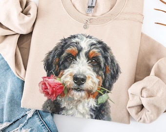 May include: Beige crewneck sweatshirt featuring a watercolor illustration of a Bernese Mountain Dog holding a red rose in its mouth. A delicate silver necklace rests on the sweatshirt. The sweatshirt is paired with distressed blue jeans.