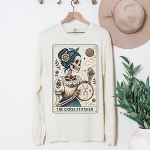 May include: A cream-colored long-sleeve shirt featuring a graphic of a skeleton with a blue hair bow and floral accents, holding a needle and embroidery hoop. The design includes yarn, flowers, and the text THE CROSS STITCHER in a rectangular frame.