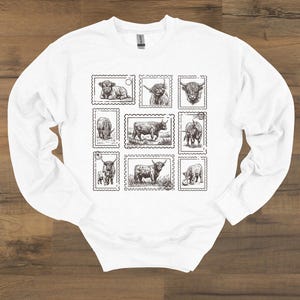 May include: White long-sleeve sweatshirt featuring a collection of Highland cow illustrations designed as postage stamps. The black and white drawings depict various poses of the cows, creating a unique and artistic design.
