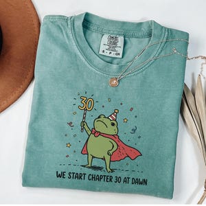 May include: A teal Comfort Colors t-shirt with a cartoon frog wearing a cape and party hat, holding a "30" sign. The text "WE START CHAPTER 30 AT DAWN" is printed below the frog. A gold necklace is draped across the shirt.