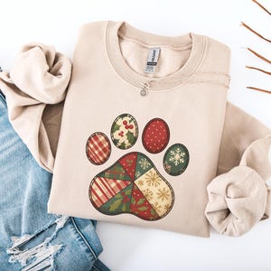 May include: A beige sweatshirt featuring a decorative paw print design. The paw print is composed of various festive patterns, including plaid, polka dots, holly, and snowflakes in red, green, and gold. A delicate silver necklace with a small pendant rests on the sweatshirt.