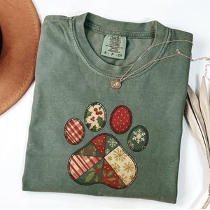 May include: A sage green Comfort Colors t-shirt featuring a patchwork paw print design. The paw print includes red, green, and white patterns with holly, snowflakes, and plaid. A gold necklace with a pendant rests on the shirt.