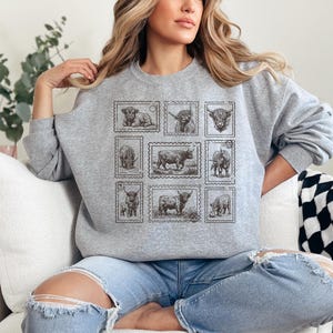 May include: A gray sweatshirt featuring a collection of nine postage stamp designs, each showcasing a different illustration of a cow. The stamps are arranged in a grid pattern on the front of the sweatshirt. The sweatshirt has long sleeves and a crew neck.