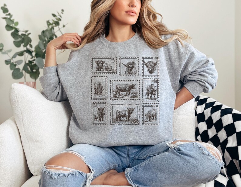 May include: A gray sweatshirt featuring a collection of nine postage stamp designs, each showcasing a different illustration of a cow. The stamps are arranged in a grid pattern on the front of the sweatshirt. The sweatshirt has long sleeves and a crew neck.