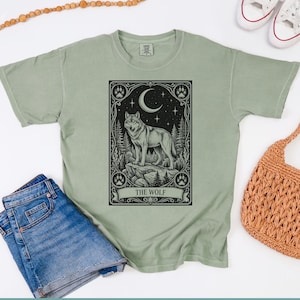 May include: A sage green short-sleeve t-shirt featuring a black graphic print of a wolf standing on a rock within a rectangular frame. The design includes a crescent moon, stars, pine trees, and paw prints, with the text THE WOLF at the bottom.