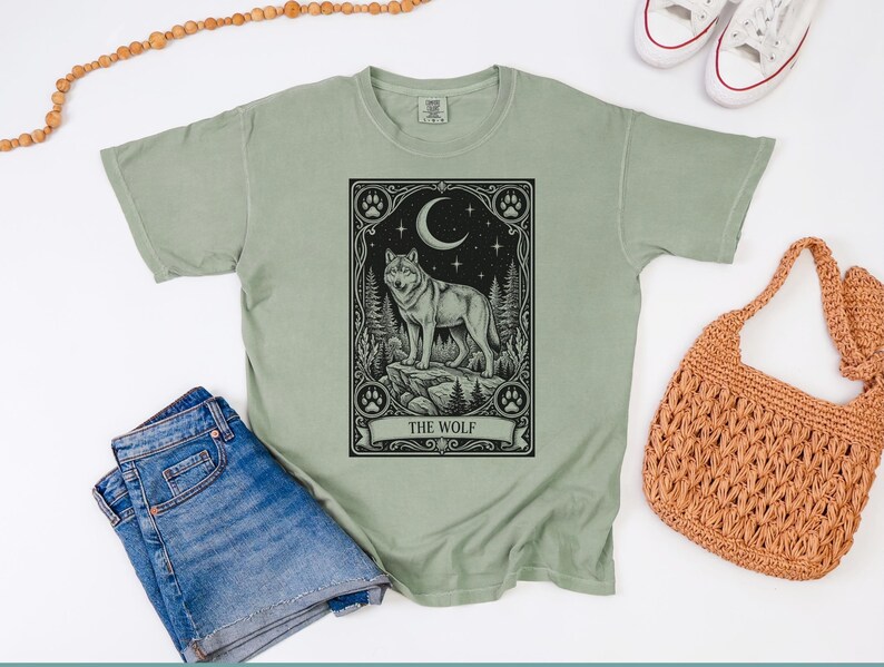 May include: A sage green short-sleeve t-shirt featuring a black graphic print of a wolf standing on a rock within a rectangular frame. The design includes a crescent moon, stars, pine trees, and paw prints, with the text THE WOLF at the bottom.