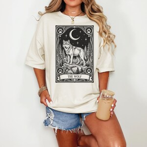 May include: A cream-colored short-sleeve t-shirt featuring a black graphic print of a wolf standing on a rock under a crescent moon and stars. The design is framed like a tarot card with paw prints in the corners and the text THE WOLF below.