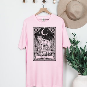 May include: A light pink t-shirt featuring a black graphic print of a wolf standing on a rock within a rectangular frame. The design includes a crescent moon, stars, pine trees, and paw prints, with the text THE WOLF printed at the bottom.