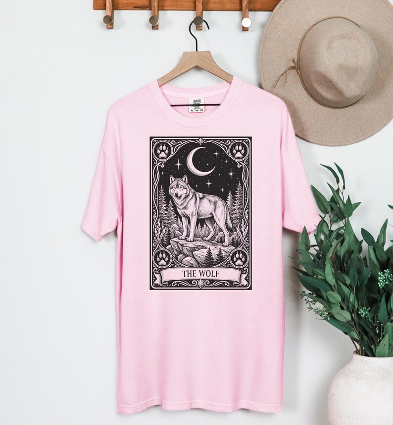 May include: A light pink t-shirt featuring a black graphic print of a wolf standing on a rock within a rectangular frame. The design includes a crescent moon, stars, pine trees, and paw prints, with the text THE WOLF printed at the bottom.