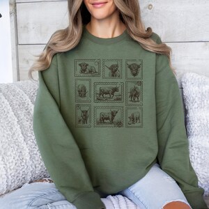 May include: An olive green sweatshirt featuring a grid of nine postage stamp-style illustrations of Highland cattle. The stamps are in a sepia tone, with detailed drawings of the animals in various poses. The sweatshirt has a crew neck and long sleeves.