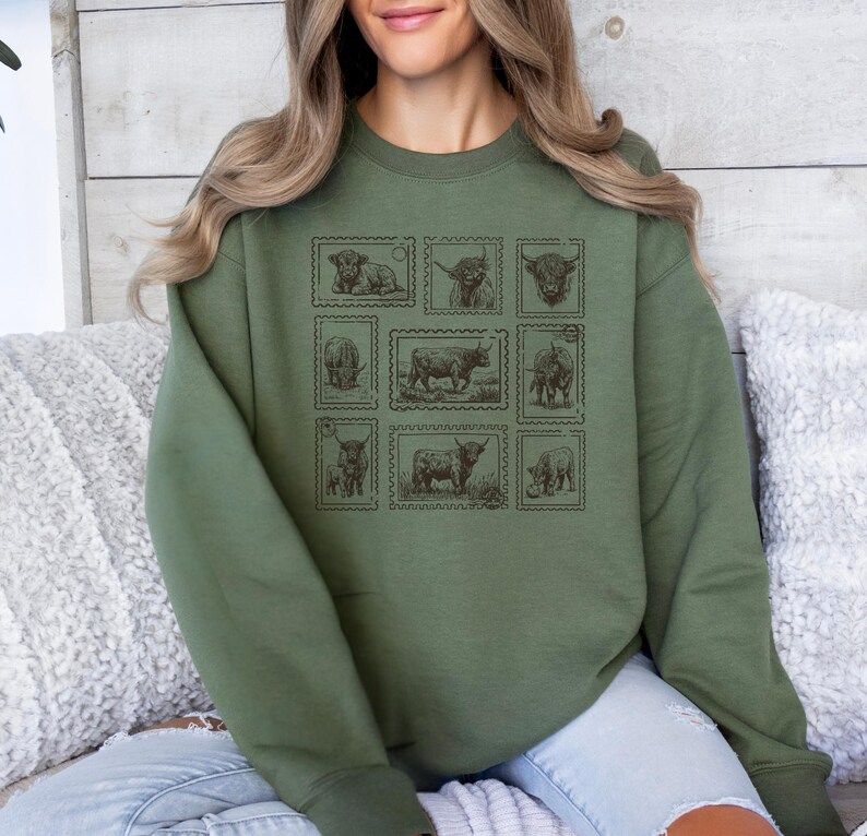 May include: An olive green sweatshirt featuring a grid of nine postage stamp-style illustrations of Highland cattle. The stamps are in a sepia tone, with detailed drawings of the animals in various poses. The sweatshirt has a crew neck and long sleeves.