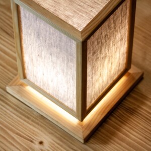 May include: A wooden cube lamp with a light linen shade. The lamp has a square base and a matching wooden frame. The light shines through the shade, creating a warm glow. The lamp is on a wooden surface.