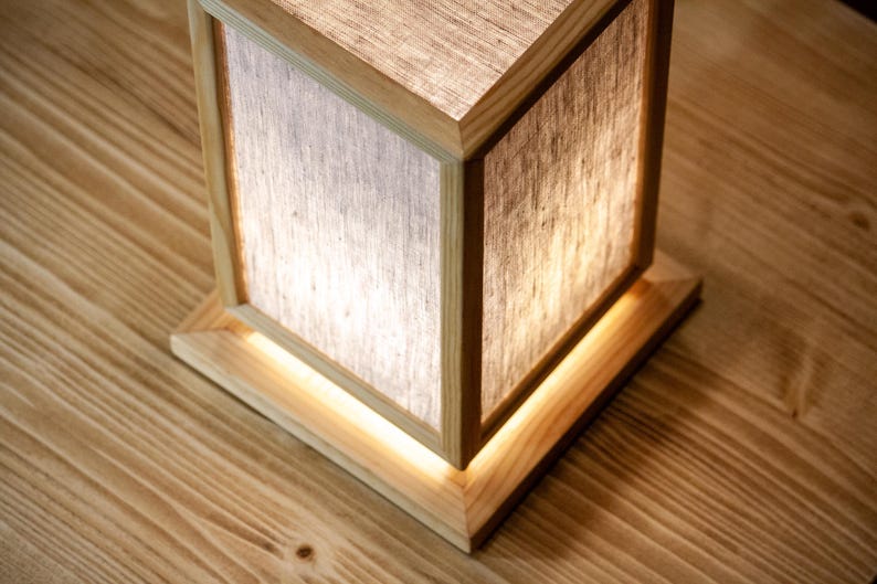 May include: A wooden cube lamp with a light linen shade. The lamp has a square base and a matching wooden frame. The light shines through the shade, creating a warm glow. The lamp is on a wooden surface.