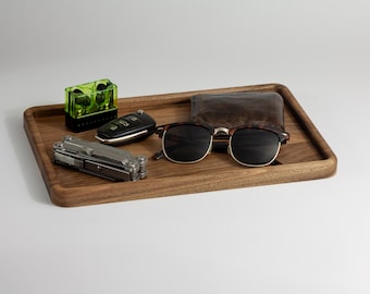 May include: A wooden tray holds various everyday carry items. These include a pair of sunglasses, a wallet, a multi-tool, a car key, and a green and black charging case. The tray is made of a light brown wood.
