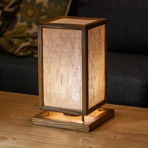 May include: A rectangular wooden table lamp with a linen shade. The lamp has a warm, inviting glow, and is set on a wooden table. The lamp's dimensions are approximately 6 inches wide, 6 inches deep, and 12 inches tall.