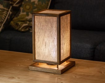 May include: A rectangular wooden table lamp with a linen shade. The lamp has a warm, inviting glow, and is set on a wooden table. The lamp's dimensions are approximately 6 inches wide, 6 inches deep, and 12 inches tall.