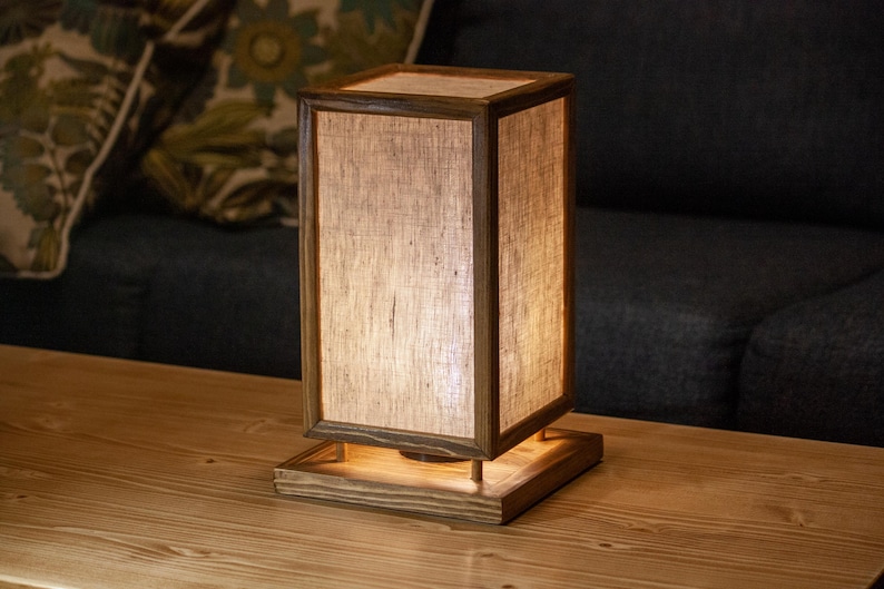 May include: A rectangular wooden table lamp with a linen shade. The lamp has a warm, inviting glow, and is set on a wooden table. The lamp's dimensions are approximately 6 inches wide, 6 inches deep, and 12 inches tall.