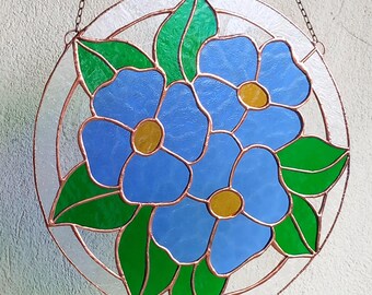 May include: A circular stained glass suncatcher featuring three blue flowers with yellow centers and green leaves, all encased in a copper foil frame. The piece is suspended by a thin metal chain, showcasing a clear glass border around the floral design.
