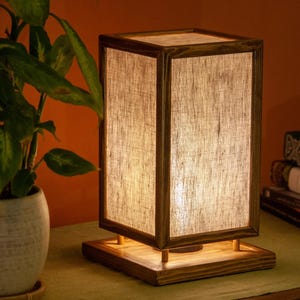 May include: A rectangular wooden table lamp with a linen shade. The lamp has a warm, inviting glow and sits on a wooden base. A potted plant and books are in the background.