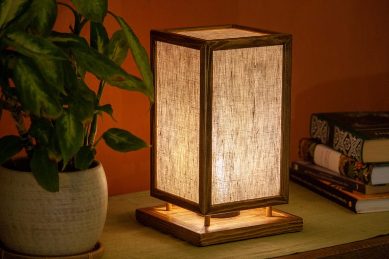 May include: A rectangular wooden table lamp with a linen shade. The lamp has a warm, inviting glow and sits on a wooden base. A potted plant and books are in the background.