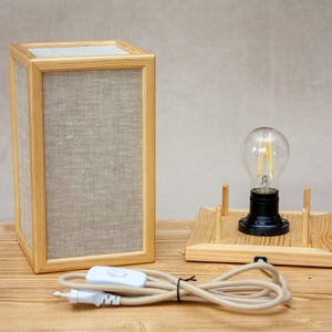 May include: A rectangular wooden table lamp with a linen shade and a light bulb. The lamp has a natural wood frame and a neutral-colored shade. A separate wooden base holds a light bulb with a black base. The lamp is connected to a power cord.