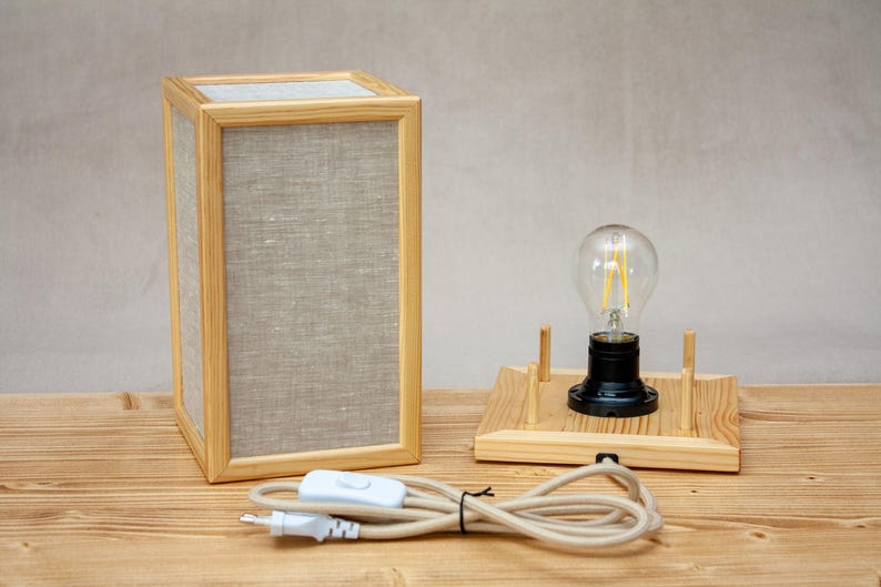 May include: A rectangular wooden table lamp with a linen shade and a light bulb. The lamp has a natural wood frame and a neutral-colored shade. A separate wooden base holds a light bulb with a black base. The lamp is connected to a power cord.