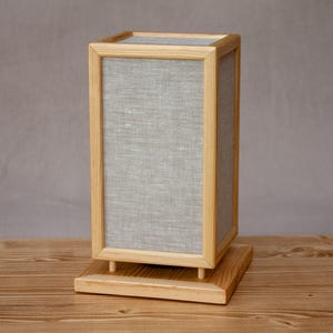 May include: A rectangular wooden lamp with a light gray linen shade. The lamp has a natural wood frame and base. The lamp is designed with a minimalist aesthetic and sits on a wooden surface.