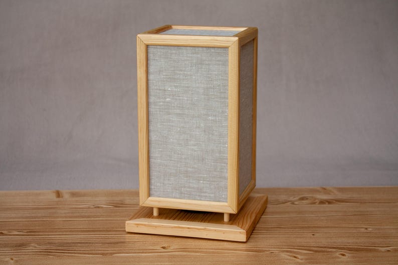 May include: A rectangular wooden lamp with a light gray linen shade. The lamp has a natural wood frame and base. The lamp is designed with a minimalist aesthetic and sits on a wooden surface.