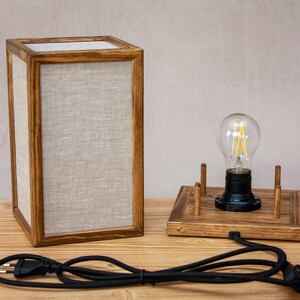 May include: A wooden table lamp with a rectangular frame and linen panels. A separate wooden base holds a lightbulb. The lamp has a black power cord and plug. The lamp is on a wooden surface.