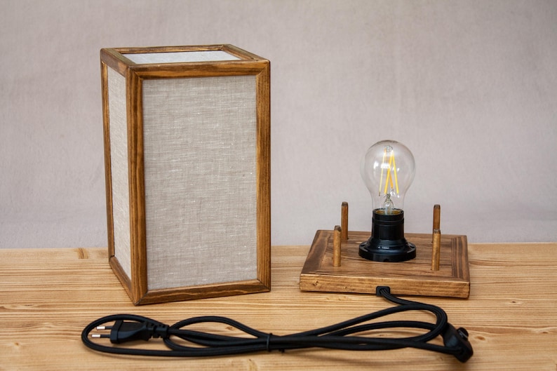 May include: A wooden table lamp with a rectangular frame and linen panels. A separate wooden base holds a lightbulb. The lamp has a black power cord and plug. The lamp is on a wooden surface.