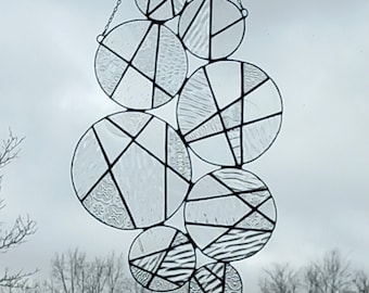 May include: A clear glass suncatcher with a series of overlapping circles, each divided by black lines. The circles have various textured patterns. The suncatcher hangs from a chain, with a cloudy sky and bare trees in the background.