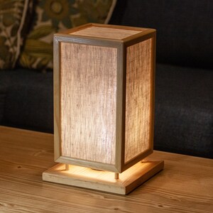 May include: A rectangular wooden table lamp with a light-colored shade. The lamp has a square base and a rectangular frame. The shade is made of a light-colored, translucent material. The lamp is sitting on a wooden table.